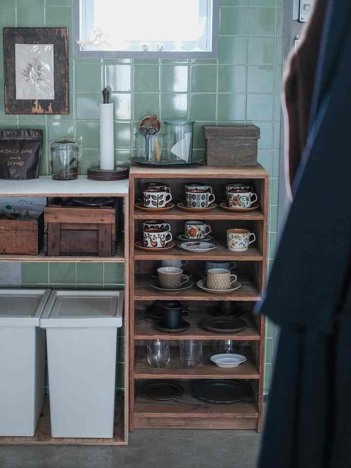 ナチュラルな木目調と淡い緑色のタイルが調和するキッチン空間、オープンシェルフに様々なコーヒーカップとキッチン用品が整頓された雰囲気のあるインテリア