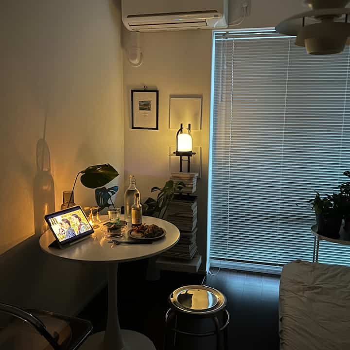 A narrow studio apartment in white and black tones featuring a round coffee table, metal stool, stacked books, and ambient lighting creating a modern atmosphere