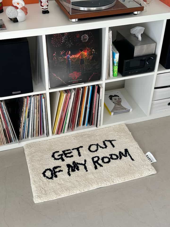 白と黒のトーンの玄関または小さな部屋、'GET OUT OF MY ROOM'のメッセージがある白いラグと本棚が配置された印象的な空間