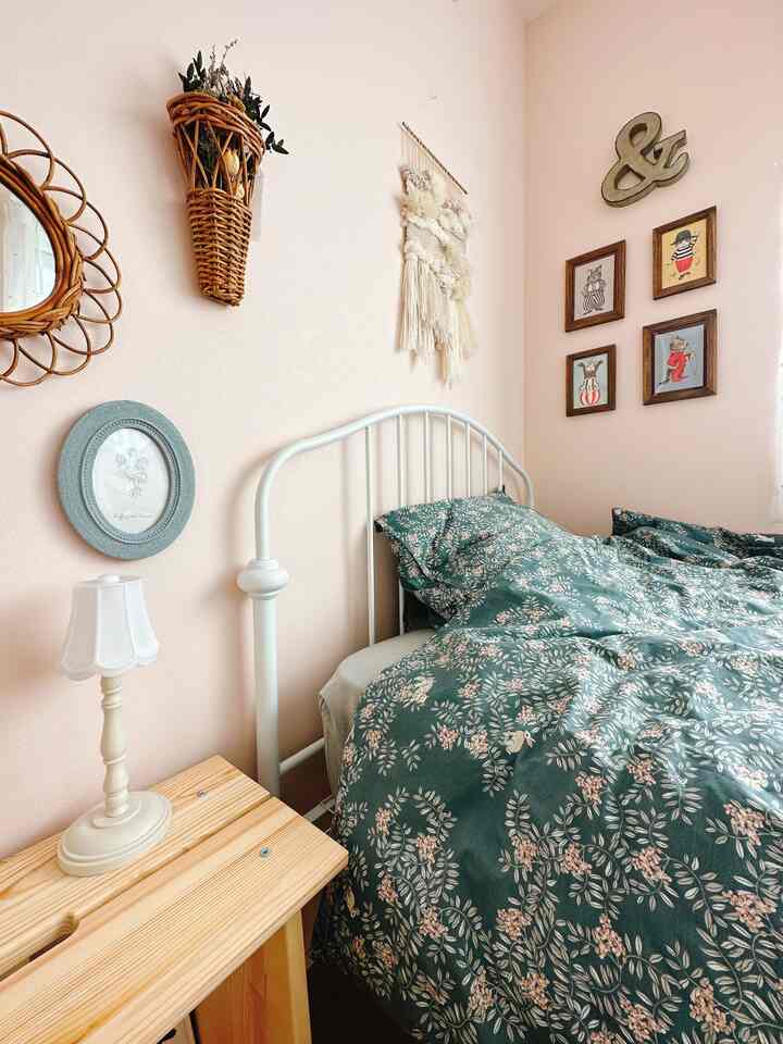パステルピンクの壁にナチュラルな木製サイドテーブルと花柄の寝具が配置された子供部屋のインテリア