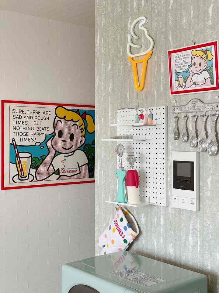 白い壁とグレーのコンクリート調の壁があるキッチンコーナー、オサムグッズのポスターとネオンのアイスクリーム照明、キッチン用品が配置された印象的な空間
