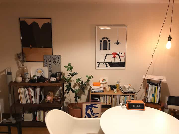 Warm natural tone living room featuring multiple bookshelves, plants, and interior posters creating a cozy atmosphere
