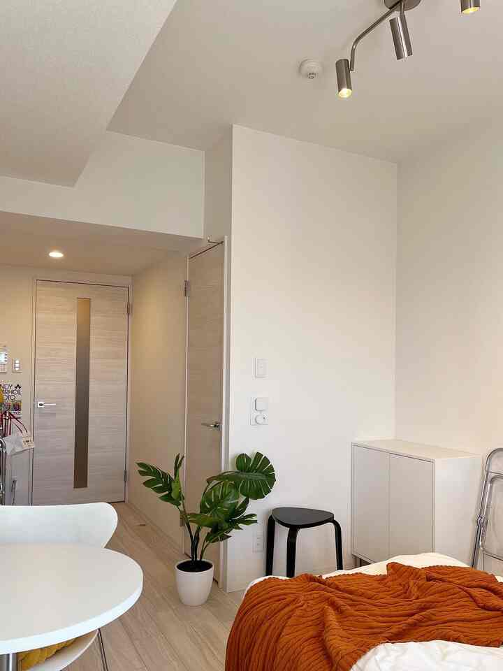 白と茶色のトーンのL字型8畳の部屋、白いダイニングテーブルとスツール、大きな観葉植物が配置された清潔感のある空間