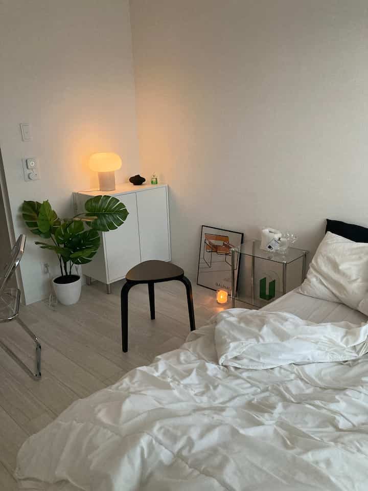 White and natural toned L-shaped bedroom featuring cabinet, plant, and transparent side table creating a clean atmosphere