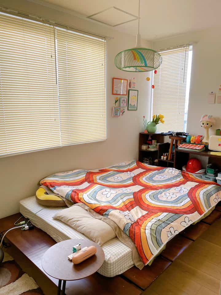 Bedroom with white walls and wood flooring, featuring a bright orange and rainbow-patterned blanket spread on the bed