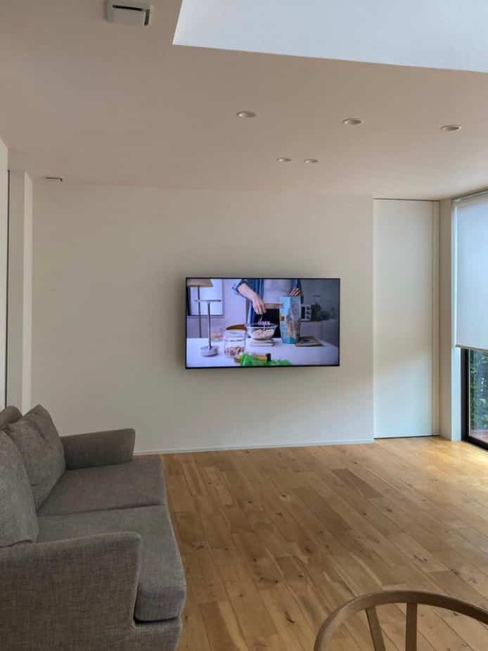 白い壁と木目調の床が印象的なシンプルなリビング、グレーのソファと壁掛けテレビが配置された空間