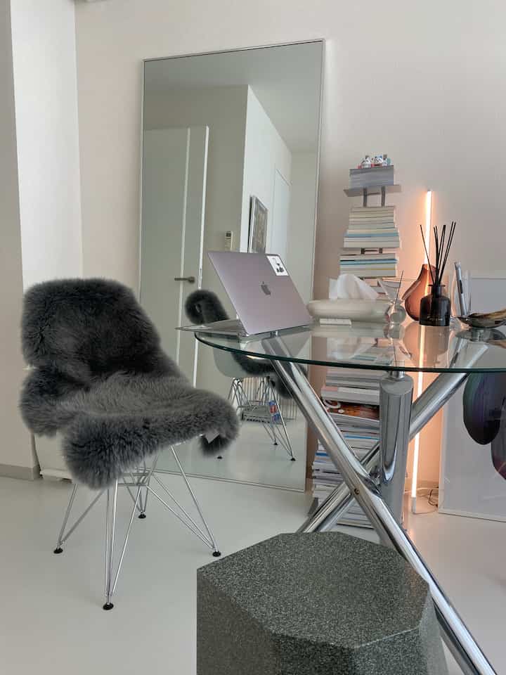 Gray and white toned entrance space featuring a large mirror, glass desk, stool, and a chair draped with gray sheepskin, creating a minimal atmosphere
