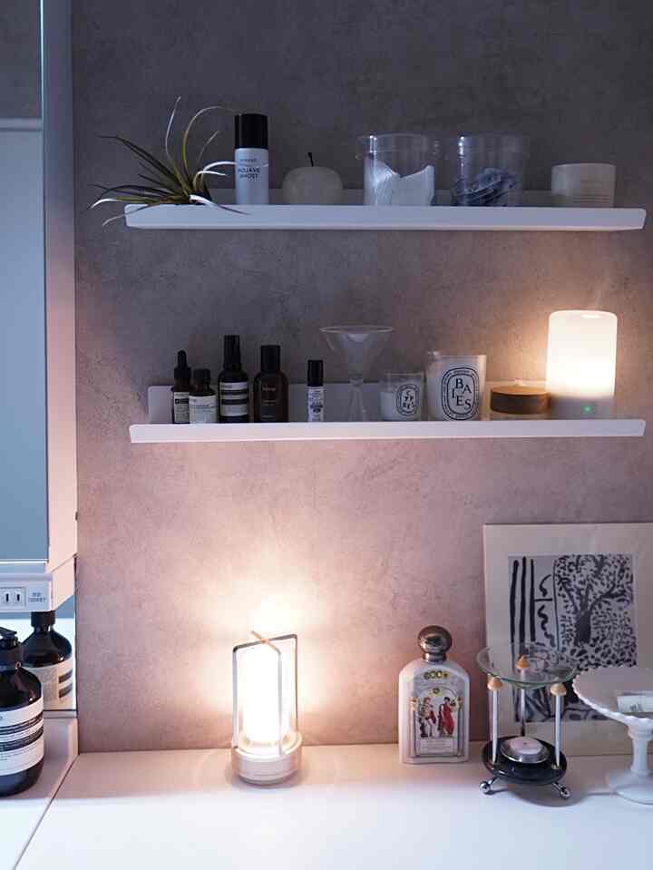 白い壁に白の棚が2段設置され、柔らかな照明とアロマディフューザー、整然としたスキンケア用品が配置された落ち着いたバスルームの空間