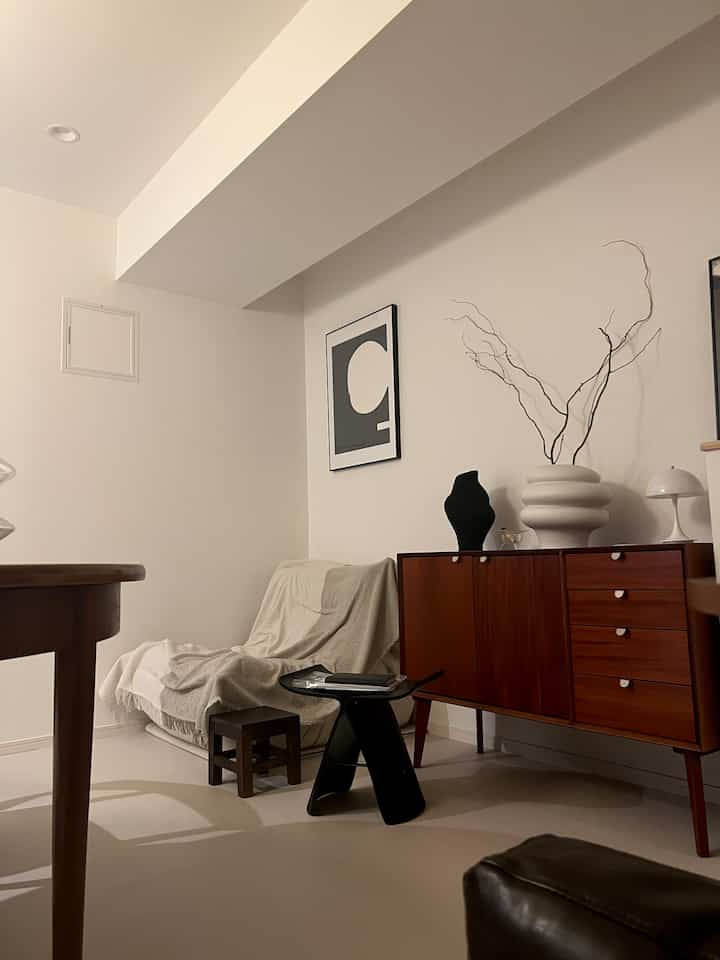 Modern kitchen and living room with natural wood tones and white walls, featuring a stool and table lamp, creating a cozy atmosphere