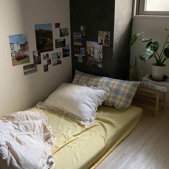 ナチュラルカラーの小さな寝室、黄色のマットレスにチェック柄と白の枕があり、壁には写真が貼られ、窓際には植物が置かれた雰囲気のある空間
