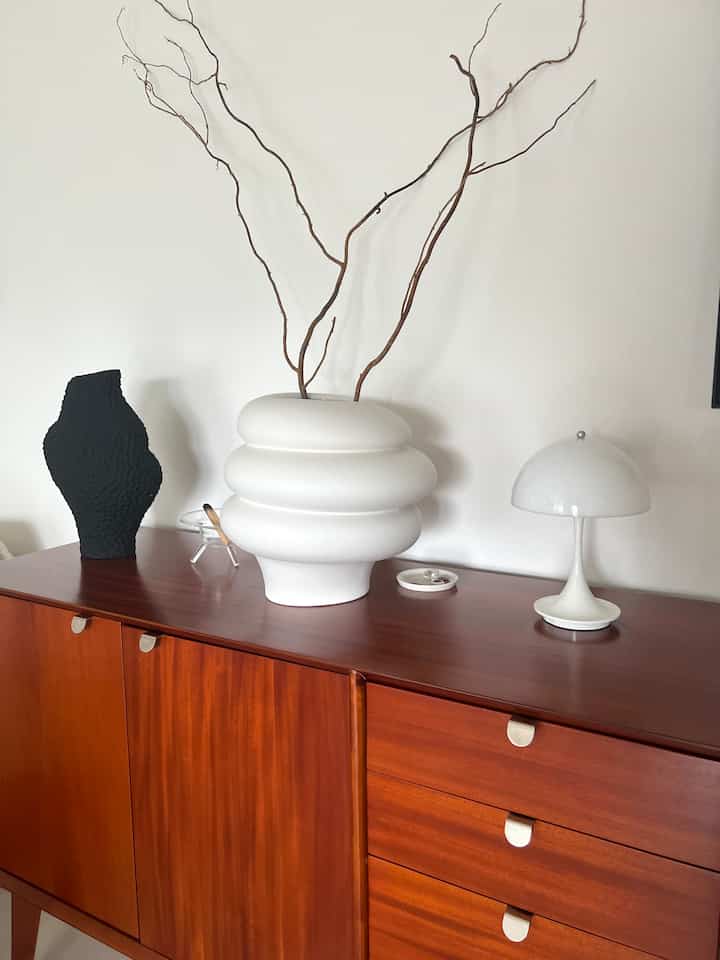 白い壁とブラウンのチェストが調和したナチュラルモダンなリビング、彫刻的な花瓶とテーブルランプが配された印象的な空間