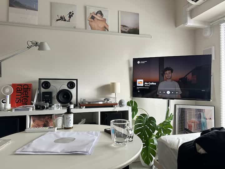 A modern living room in white and gray tones featuring a TV stand, artificial Monstera plant, and a round table with a glass mug creating a cozy atmosphere