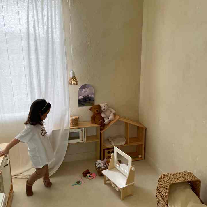 ナチュラルなベージュ色の壁と木製家具が配置された子供部屋、白いコットンカーテンとおもちゃがある雰囲気のあるインテリア