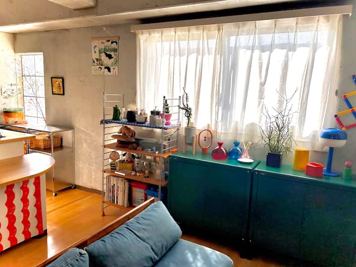Natural-toned studio apartment featuring a blue sofa, stainless steel kitchen cart, various plants, and cafe-style decor creating a cozy atmosphere