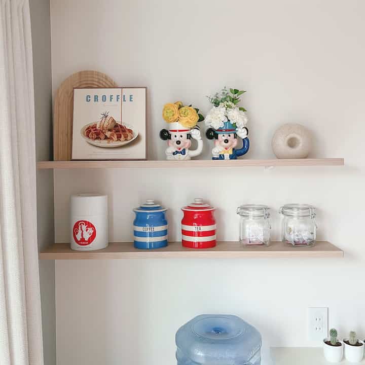 白い壁と木目調の棚が配されたダイニングスペース、ヴィンテージのミッキーマグと茶葉収納ジャーが整然と並ぶ雰囲気のあるインテリア