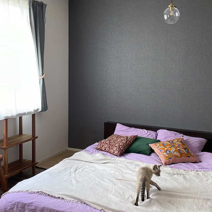 グレーの壁とベージュのブランケットがある寝室、ライトブラウンの木製棚と猫が配置されたシンプルでナチュラルなインテリア