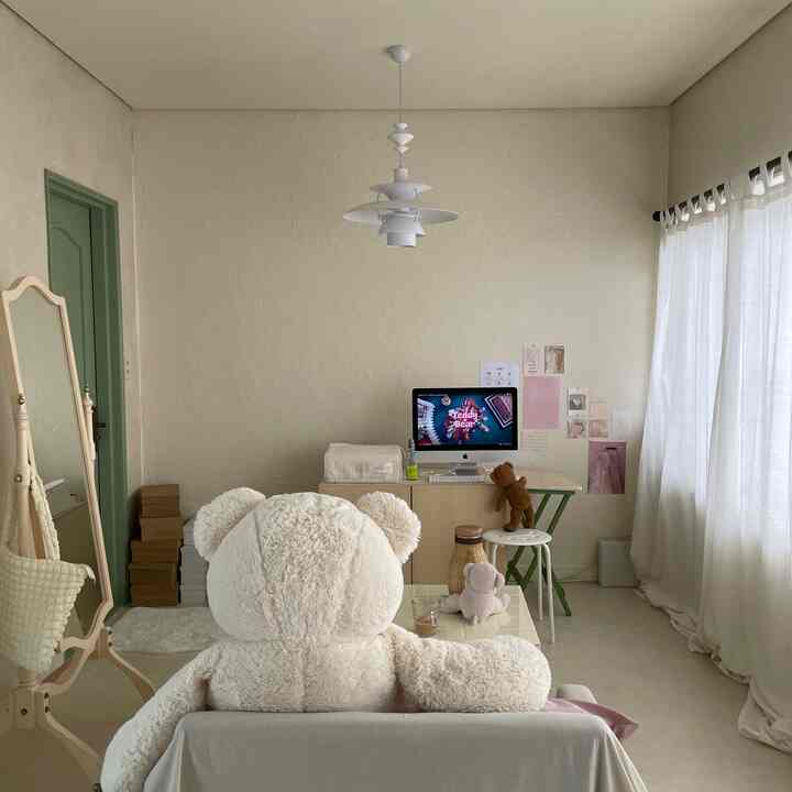 柔らかなベージュトーンのリビング、大きなテディベアがソファに座り、白いカーテンとパソコンデスクが配置されたシンプルな空間