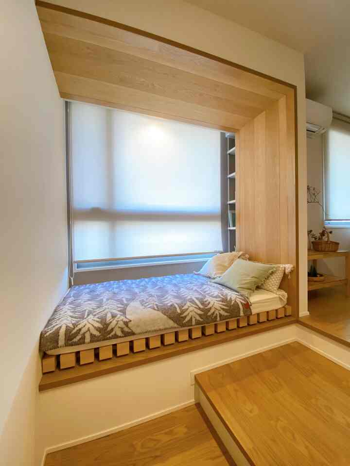 白い壁とナチュラルな木目調が調和する6畳の寝室、窓辺にくつろげる窓辺の席とブラインドが配置された印象的な空間