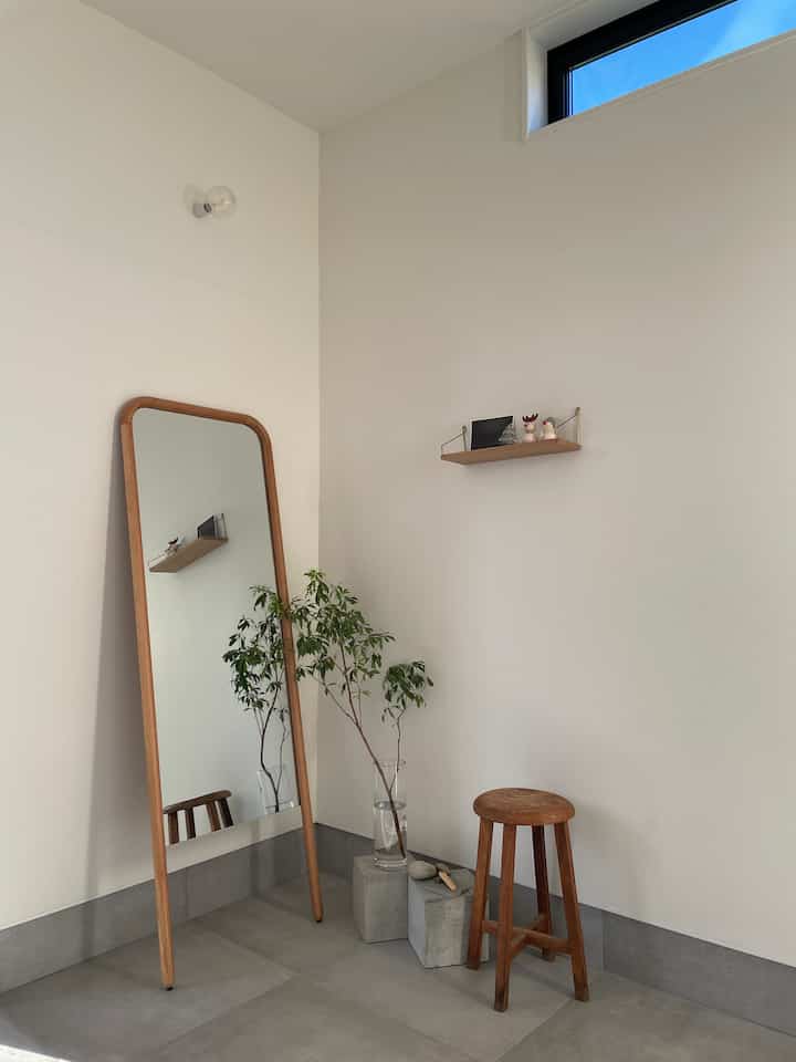 白い壁とグレーの大判タイル床の玄関スペース、大きな木製フレームの鏡と木製スツール、観葉植物が配置されたナチュラルでミニマルなインテリア