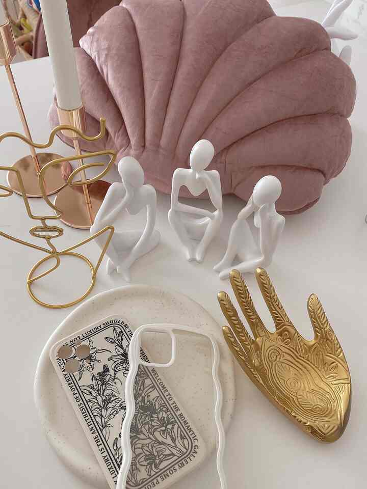 A white table featuring a pink shell-shaped cushion, gold decorative hand and candlesticks, and white abstract figurines showing a delicate decorative setup