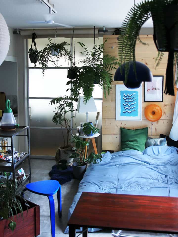 Natural toned bedroom with blue bedding, stool, and multiple plants creating a cozy atmosphere