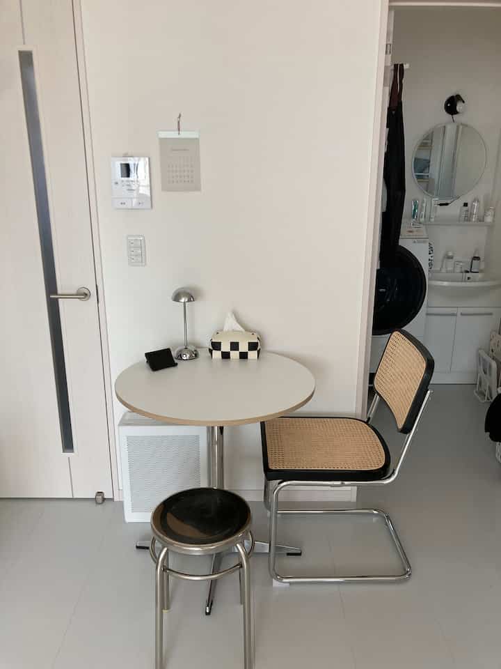 白とナチュラルカラーの狭い空間、丸テーブルとラタンチェア、ステンレススツールが配置されたミニマルな雰囲気のホームカフェ