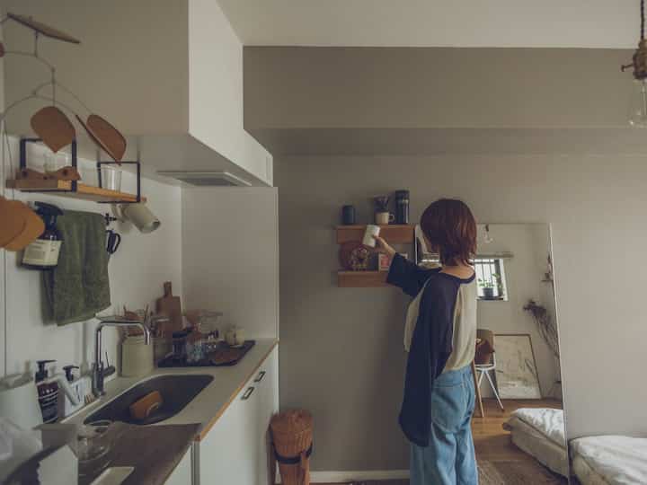 グレーのアクセントウォールと白いキッチンが調和した一人暮らしの空間、壁掛け棚とおうちカフェのアイテムが配置されたミニマルでナチュラルなインテリア