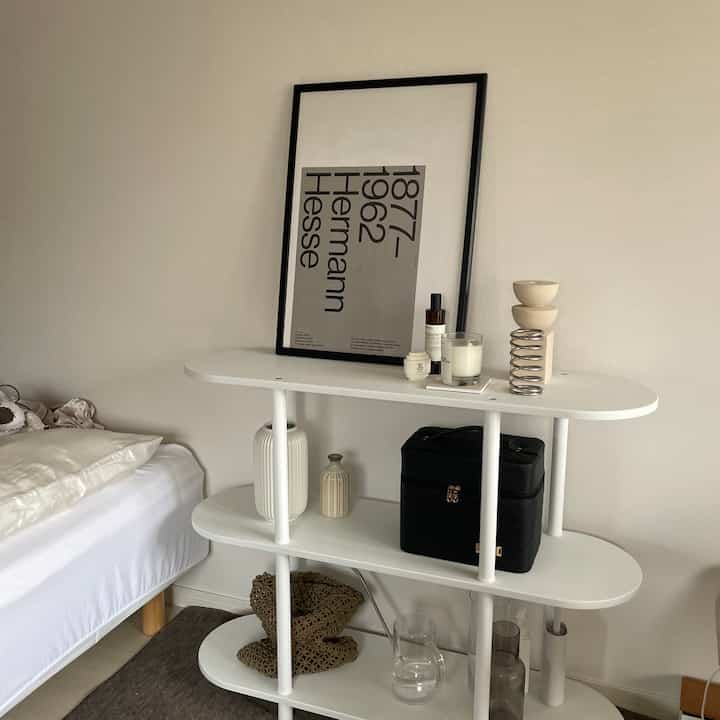 モノトーンの寝室、白い3段棚とポスターが配置されたシンプルなレイアウトの空間