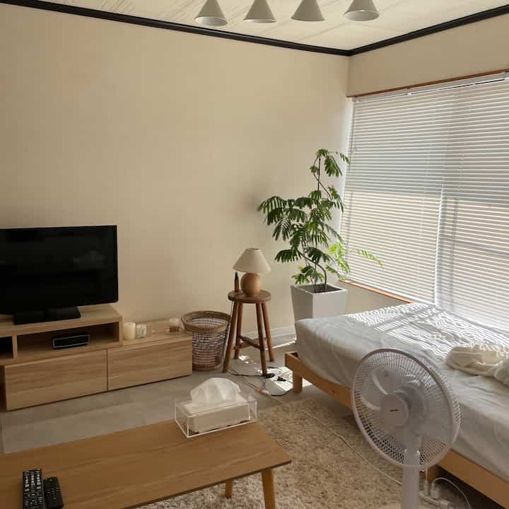 柔らかいベージュと木目調の寝室空間、ベッドとライトウッドのフレーム、ナイトテーブル、テレビスタンドが配置された落ち着いた雰囲気のインテリア