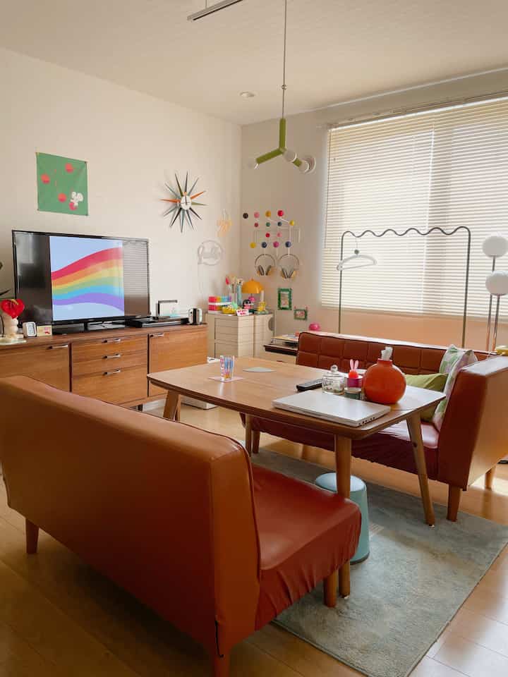 ナチュラルな木目調とブラウンソファが配置された二人暮らし向けのリビングダイニング、明るいブラインドとカラフルな小物が印象的な空間