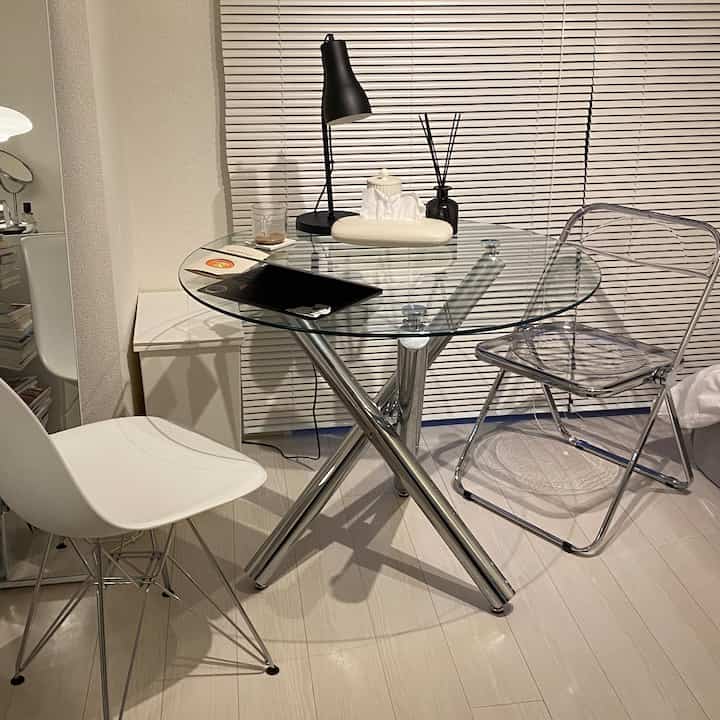 Modern, compact dining room featuring transparent glass table with chrome legs, clear chairs, and white walls with blinds creating a clean atmosphere.