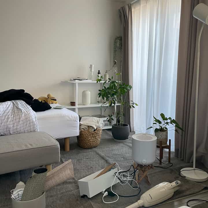 白とグレーのトーンのワンルーム、ベッドやカーペット、植物、フロアランプが配置された自然な雰囲気の空間