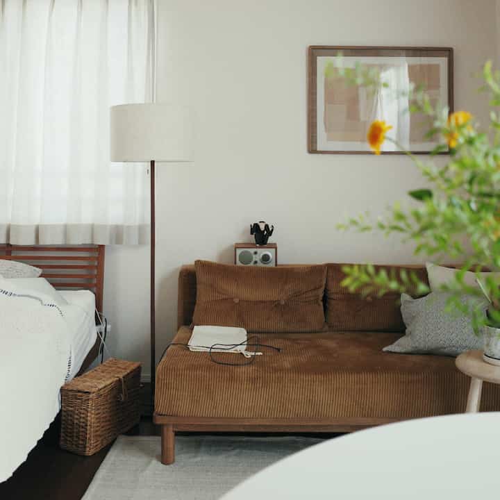 白い壁と茶色のソファがあるナチュラルな色調の8畳リビング、ベッドとフロアランプが配置された落ち着いた空間