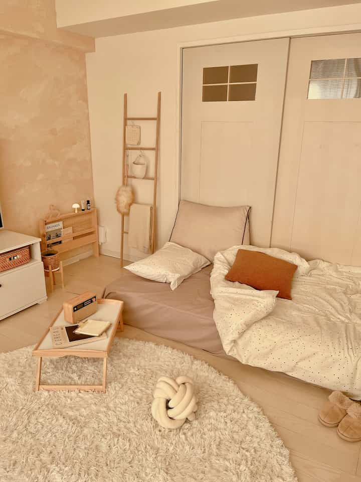 ベージュとナチュラルカラーのワンルームリビング、マットレスベッドとラグ、サイドテーブルが配置された落ち着いた空間