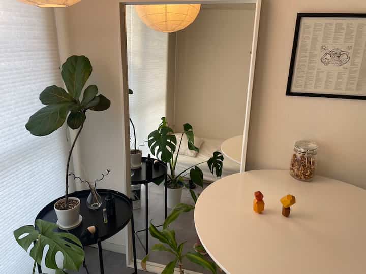 白とベージュの落ち着いた色調の小さな部屋、複数の植物と白い丸テーブル、大きな全身鏡が配置された自然な雰囲気のインテリア