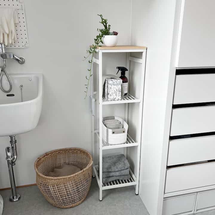 白い壁と床のバスルームに設置されたスリムな3段ラックが、タオルとバスケットを整然と収納した雰囲気のある空間