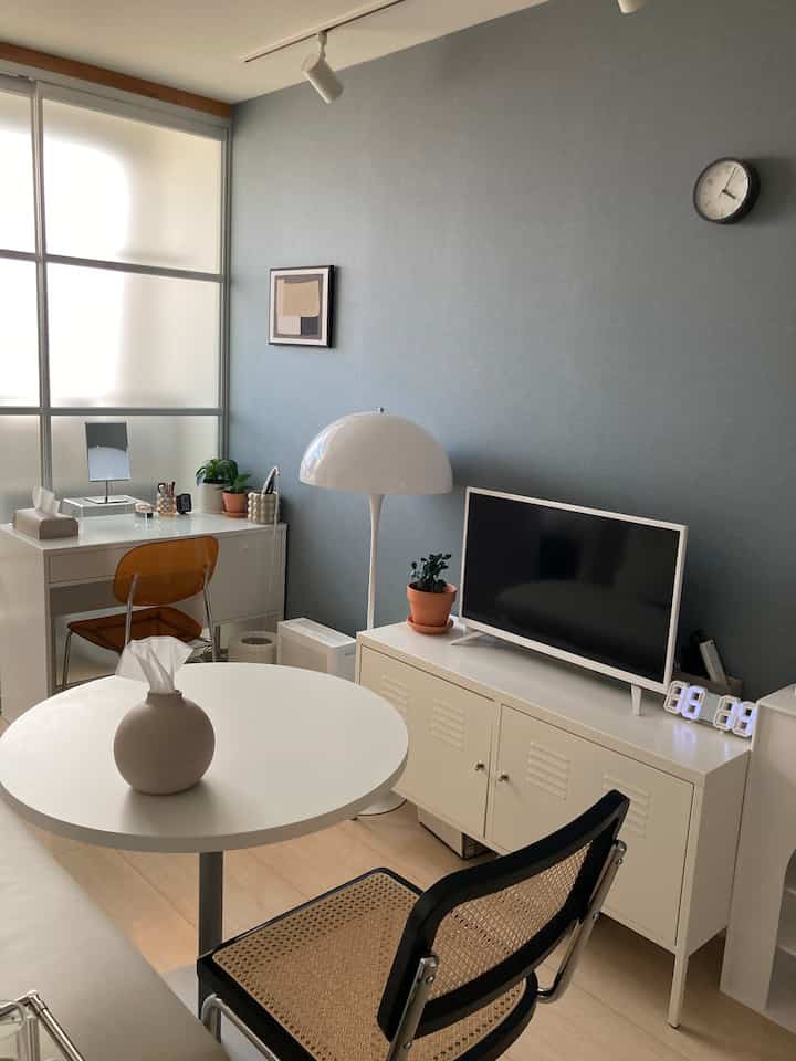 白と青のトーンが特徴的な狭いリビング、シンプルな家具とキャビネット、デスク、丸テーブルが配置された落ち着いた雰囲気のインテリア