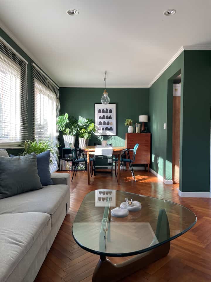 Natural modern living room with deep green walls, wooden furniture, gray sofa, glass coffee table, and indoor plants arranged invitingly