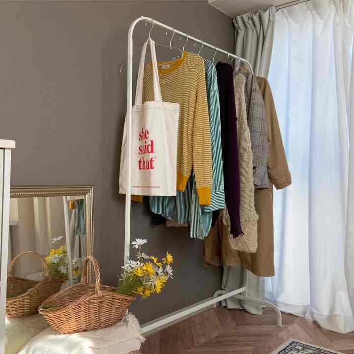 ホワイトカーテンとブラウンウッド床があるナチュラルでシンプルな狭い部屋、衣類がかかったコートラックとゴールドの縁の鏡が配置された空間