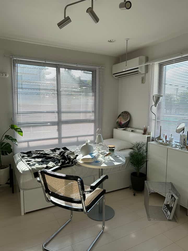 ホワイトトーンのワンルーム寝室、シンプルなベッドと丸テーブル、アームチェア、フロアランプ、植物が配置されたモダンな空間