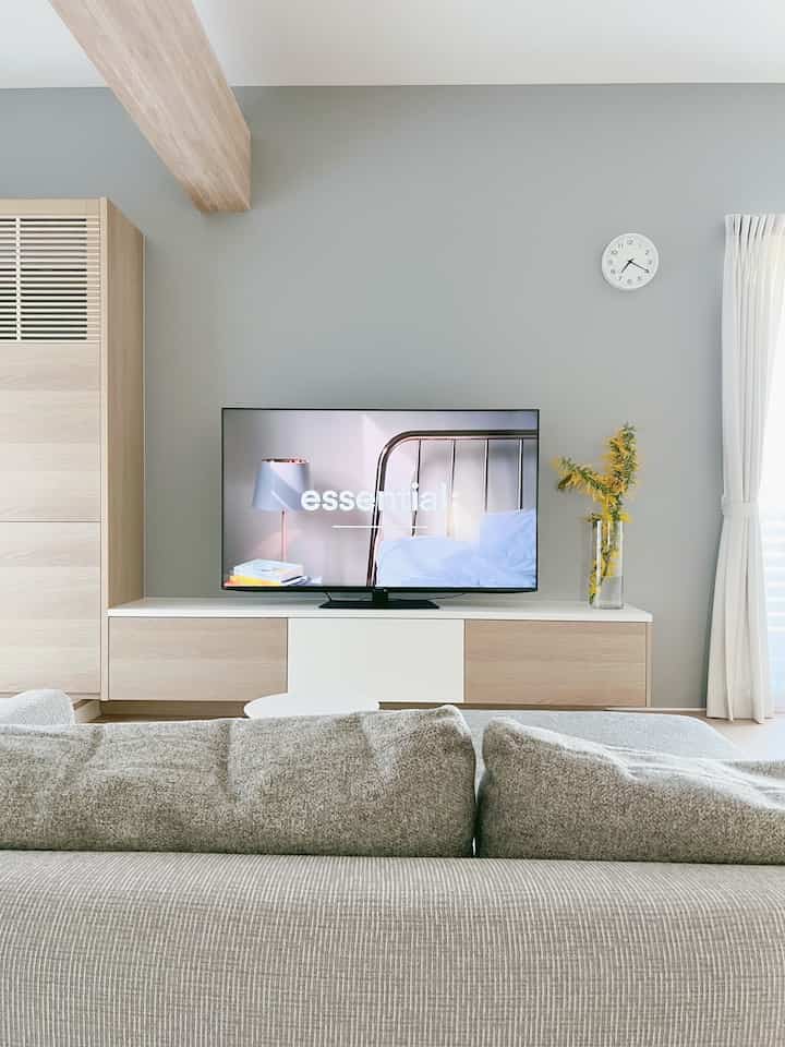 ナチュラルカラーとグレーのトーンのリビング、快適なグレーのソファと壁掛けアナログ時計、テレビ台、そして黄色いミモザが配置されたシンプルでモダンな空間