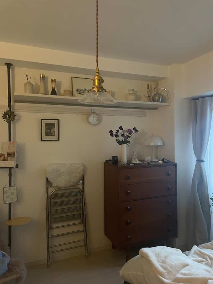 ホワイトとブラウンのナチュラルな色調の狭い部屋、ヴィンテージ小物を置いた木製チェストとステンレス製の折りたたみチェアが配置された印象的な空間