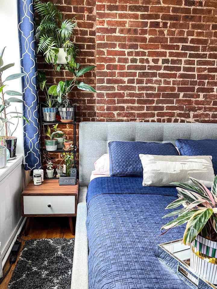 Small bedroom with dominant red brick wall and blue bedding, cozy space accented with plants and a nightstand