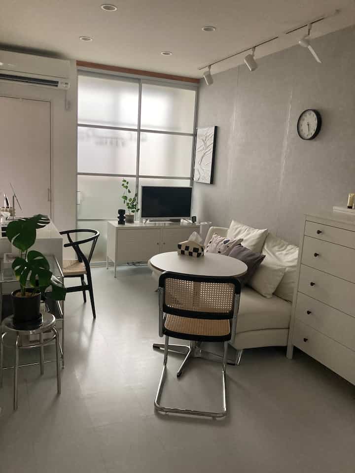 Monotone modern 1LDK living room featuring white furniture, rattan chairs, and potted plants in a simple setting