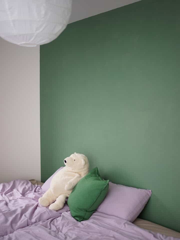 Simple bedroom with green accent wall and lavender bedding, featuring a teddy bear indicating a kid-friendly space