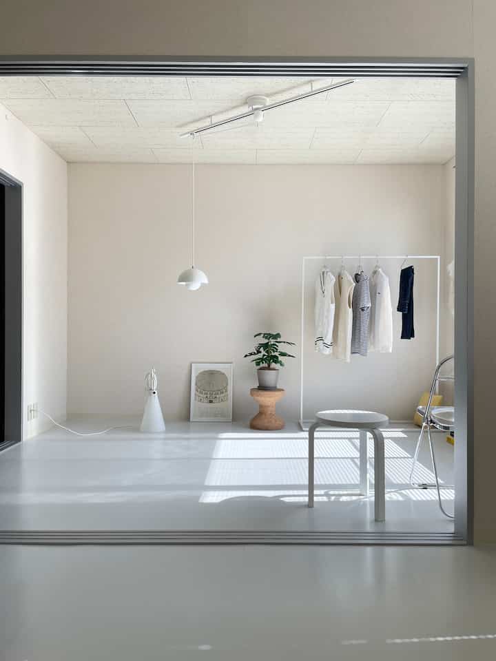 A simple studio apartment with white and natural tones, featuring a stool and clothing rack in a bright, clean interior