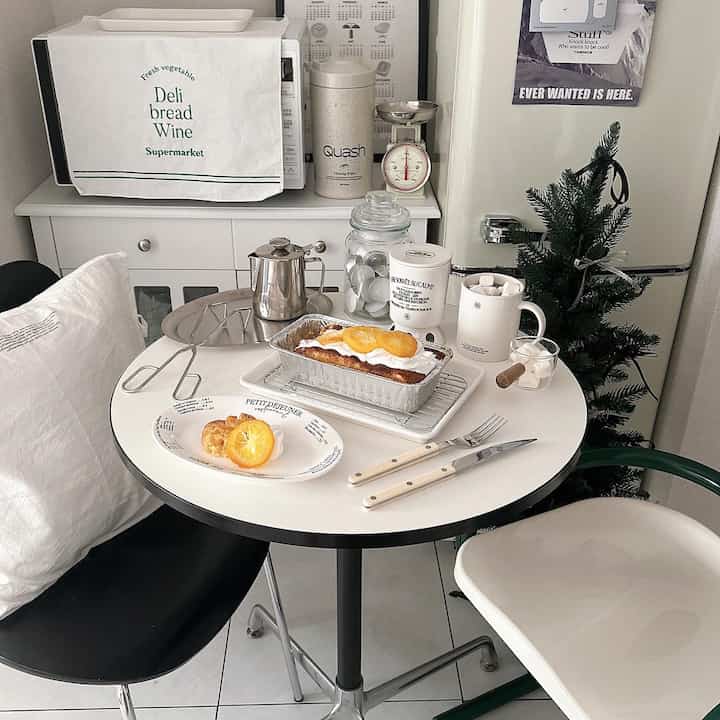 A cozy home cafe space features a small white and black dining table with coffee and orange pound cake in a compact kitchen area