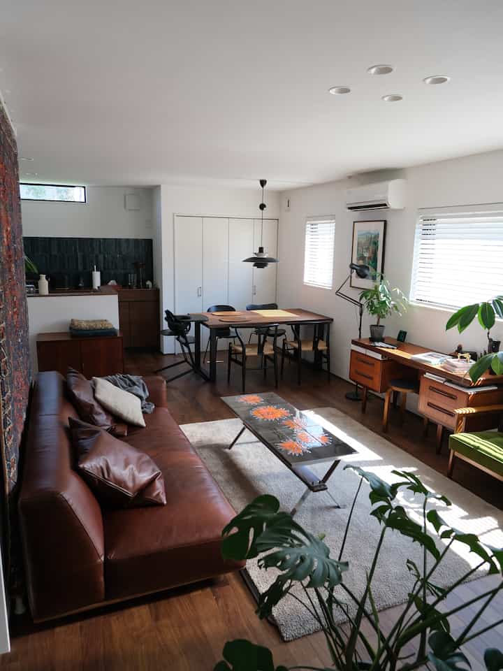 Natural modern living and dining room with brown wood tones and white walls, featuring brown leather sofa, wooden dining table, plants, and pendant light