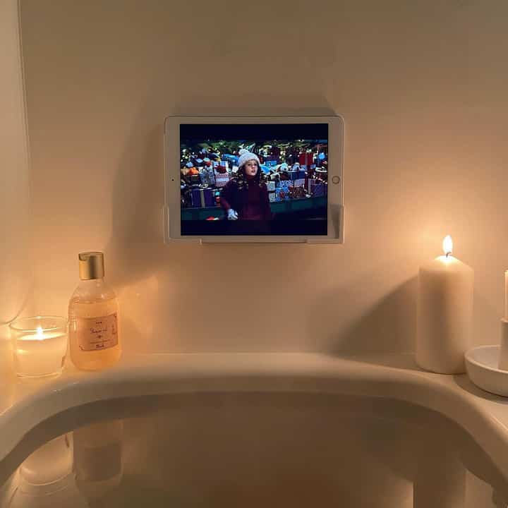 白を基調とした浴室、浴槽の縁にキャンドルが置かれ、壁に掛けられたタブレットがある居心地の良いリラクゼーション空間