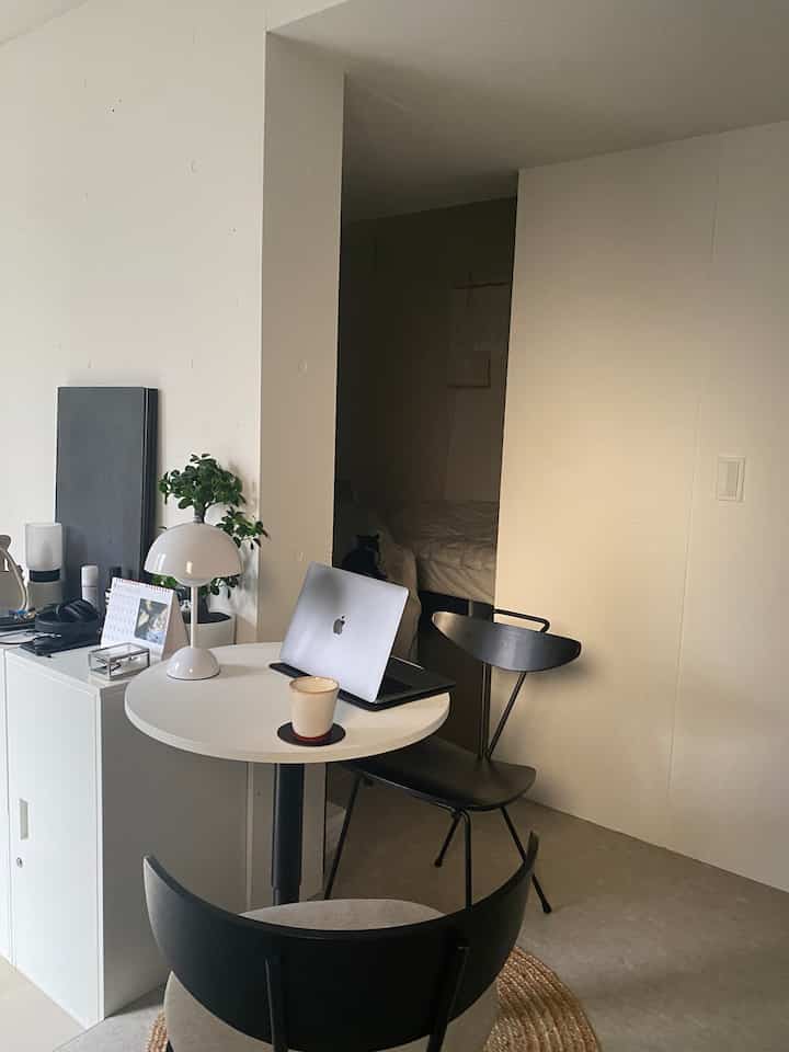 A narrow, simple single household home office space dominated by white and black tones, featuring a small round table, chairs, laptop, bedroom in background, and a cat on the bed.
